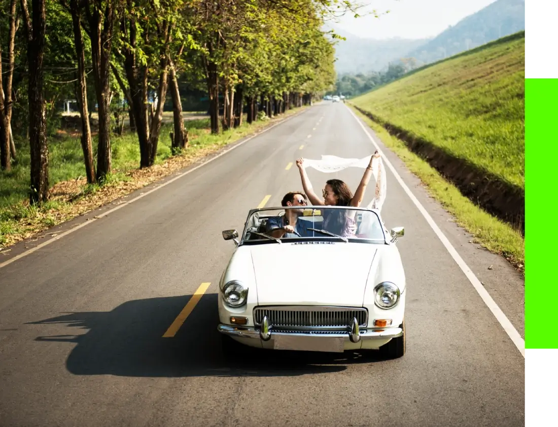 Two people driving a white convertible on a tree-lined road, holding a white cloth above their heads.