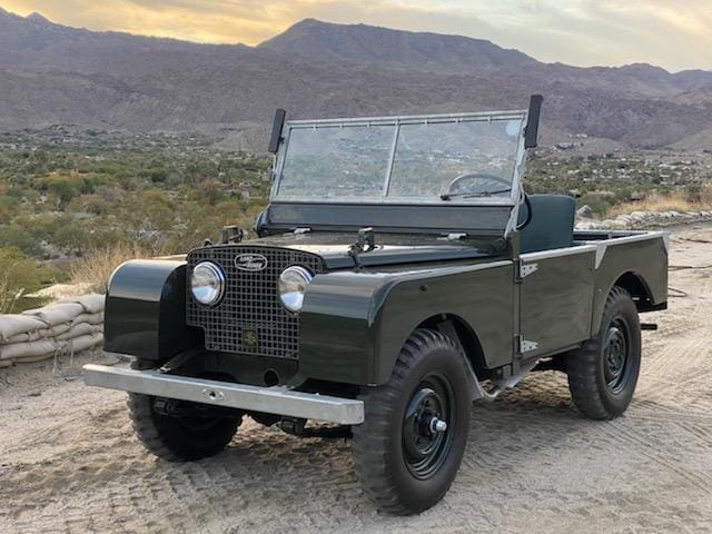 Classic green off-road vehicle parked on a dirt path with mountains in the background.