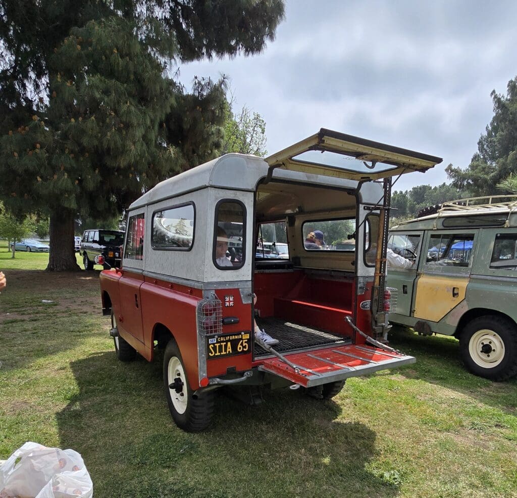 Vintage red and white Land Rover with open rear door at an outdoor event.