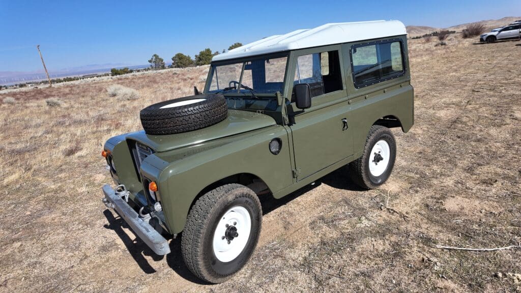 Classic olive green Land Rover parked on dry grass under clear sky.