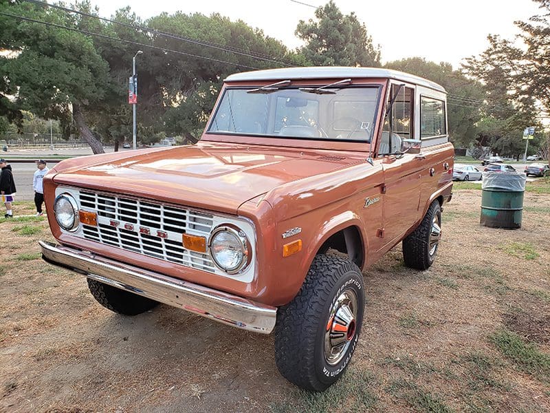 Classic orange Ford Bronco parked on grass near trees.