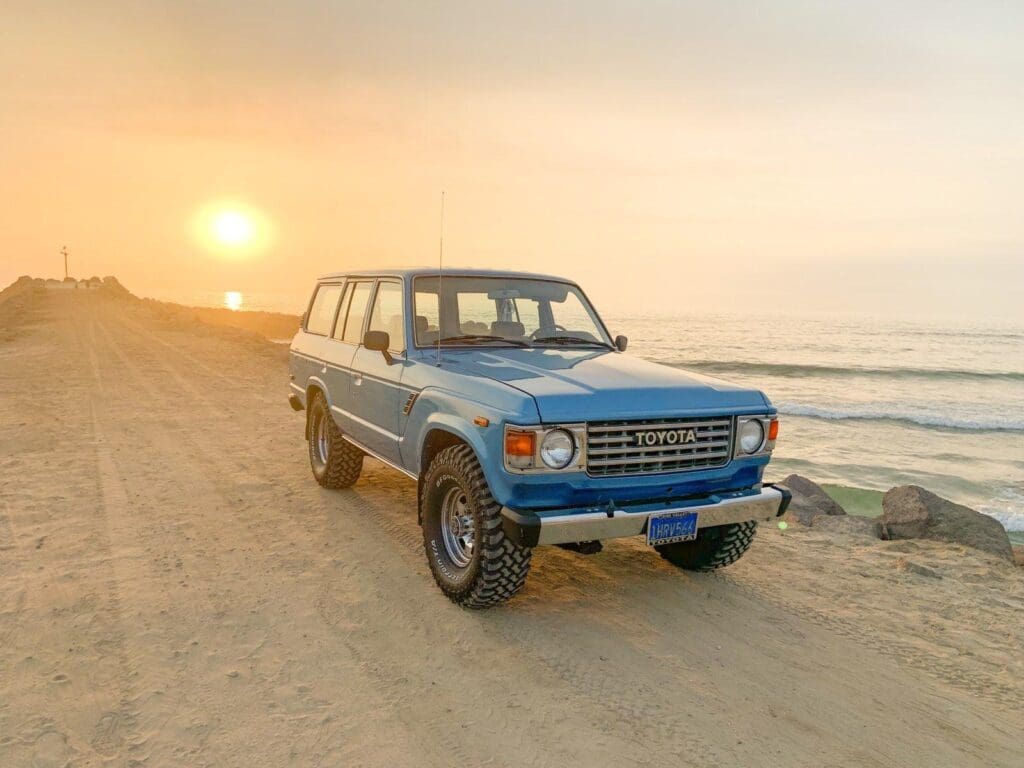 Classic blue SUV parked on a sandy beach at sunset.