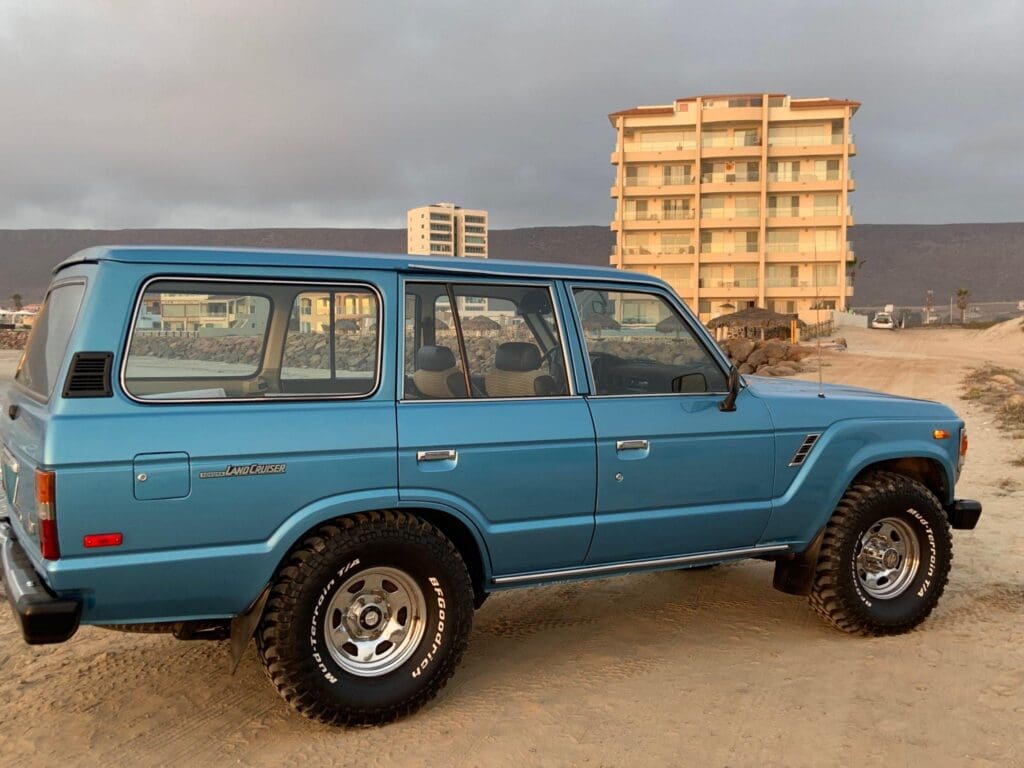 Classic blue SUV parked near a sandy beach with buildings in the background.