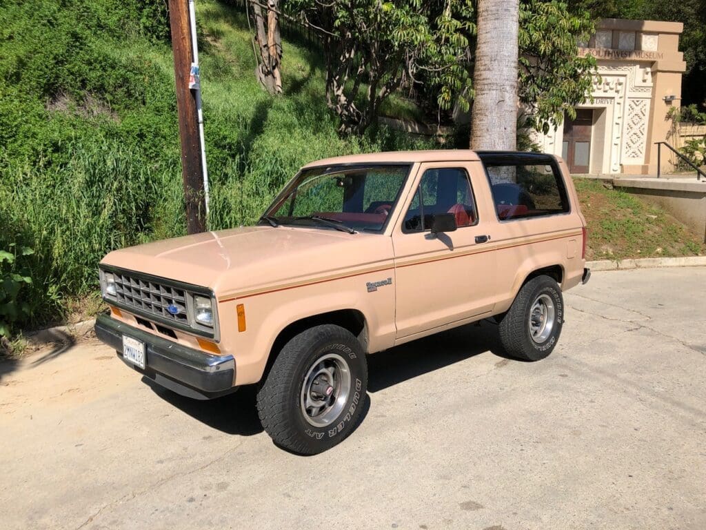 A beige vintage SUV parked on a street with greenery in the background.