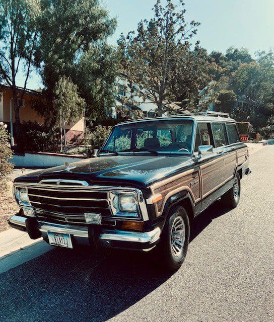 Classic black Jeep Wagoneer parked on a sunny street with trees in the background.