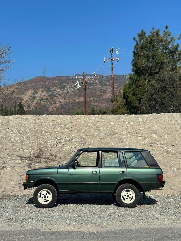 Green SUV parked on a gravel road near a hill and trees.