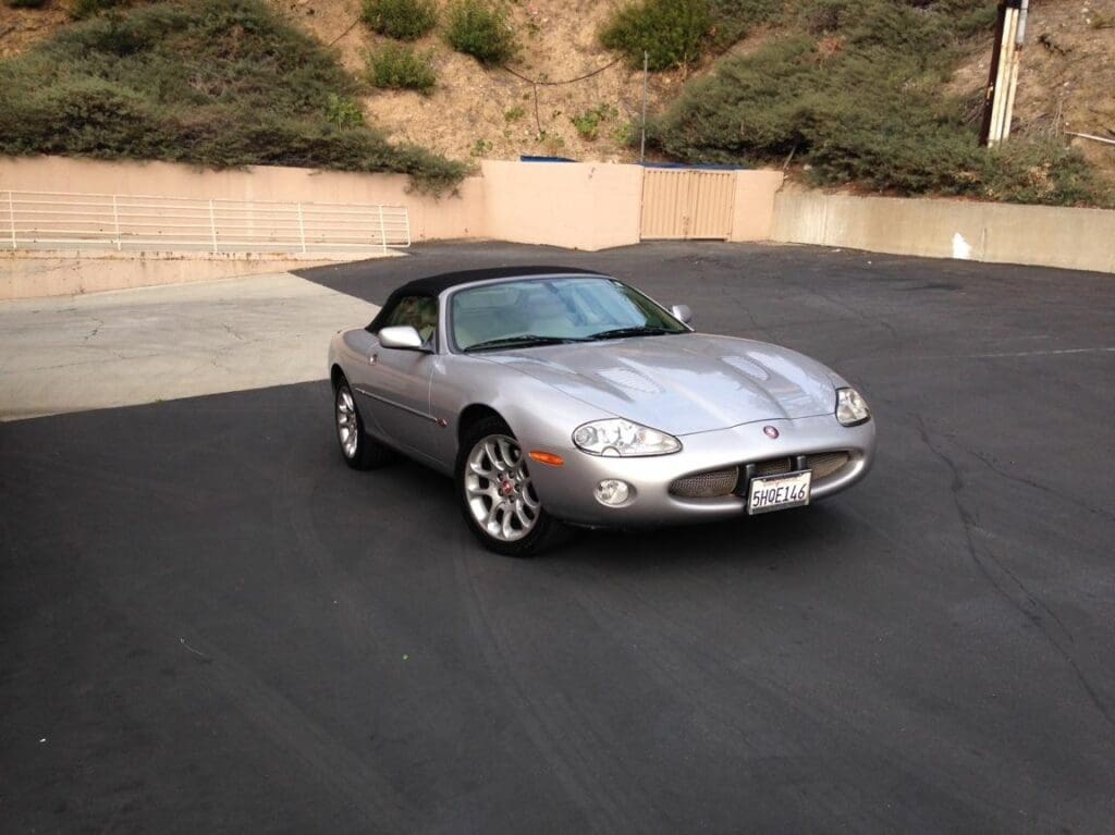 Silver Jaguar XK8 convertible parked on asphalt with hills in background.