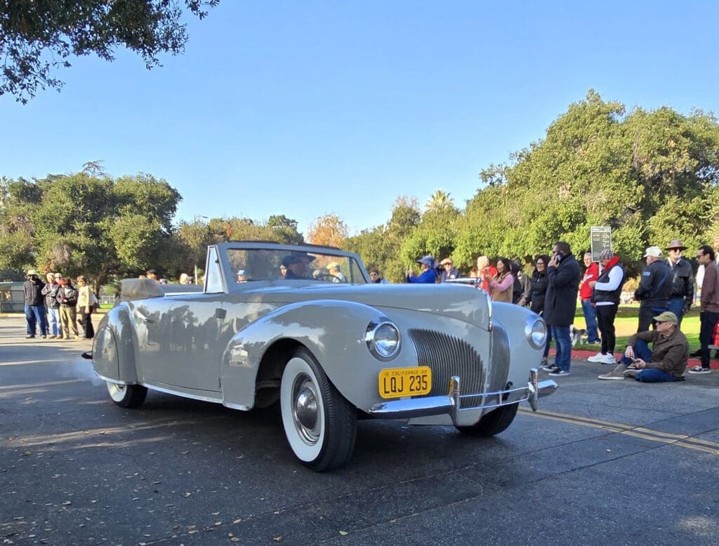Classic silver convertible car parked outdoors with people nearby.