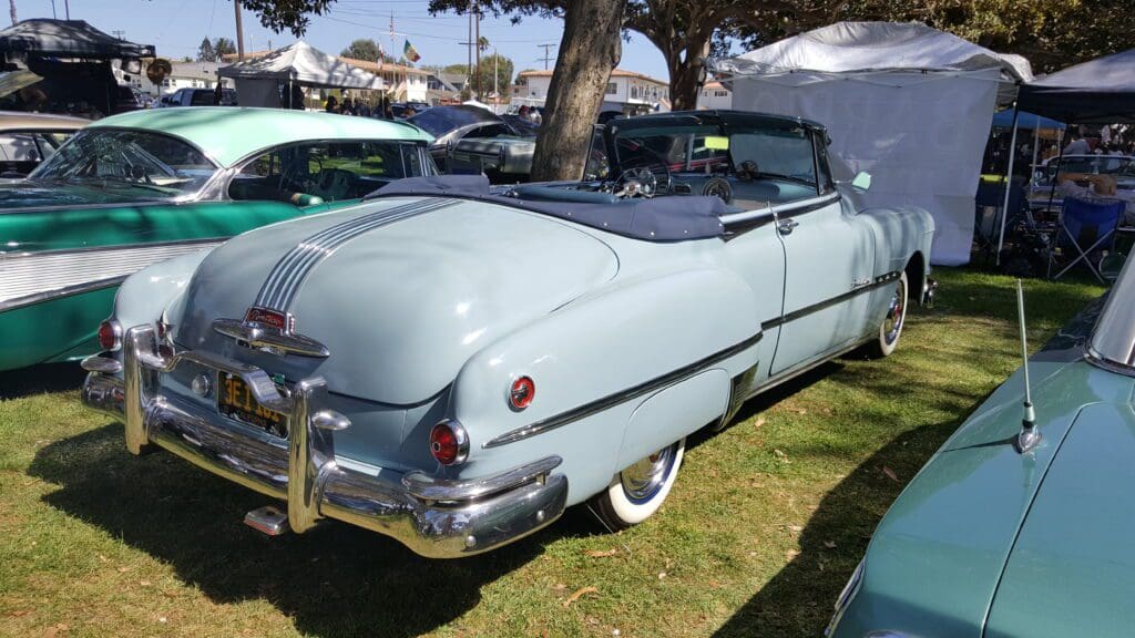 Classic light blue convertible car parked under a tree at an outdoor event.