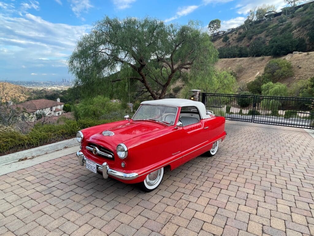 Classic red vintage convertible car parked on a stone driveway under a tree.