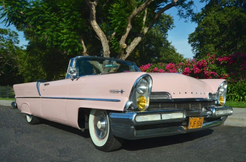 Vintage pink convertible car parked outdoors with trees and flowers.