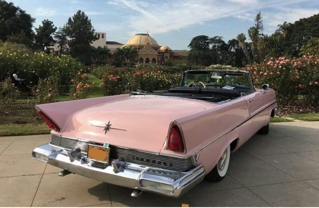 Classic pink convertible car parked near rose gardens with a dome building in the background.