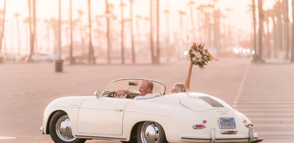 Couple drives in a white convertible with a bouquet held up joyfully.