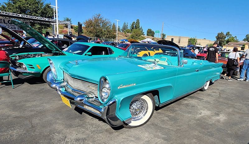 Classic turquoise convertible car with whitewall tires at a sunny car show.
