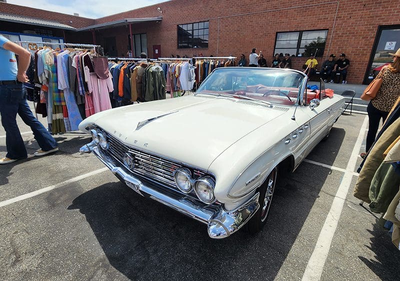 A vintage white convertible car parked near clothing racks outdoors.
