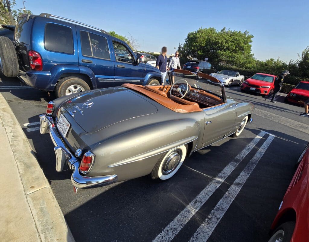 Classic silver convertible car parked in a lot with people nearby.