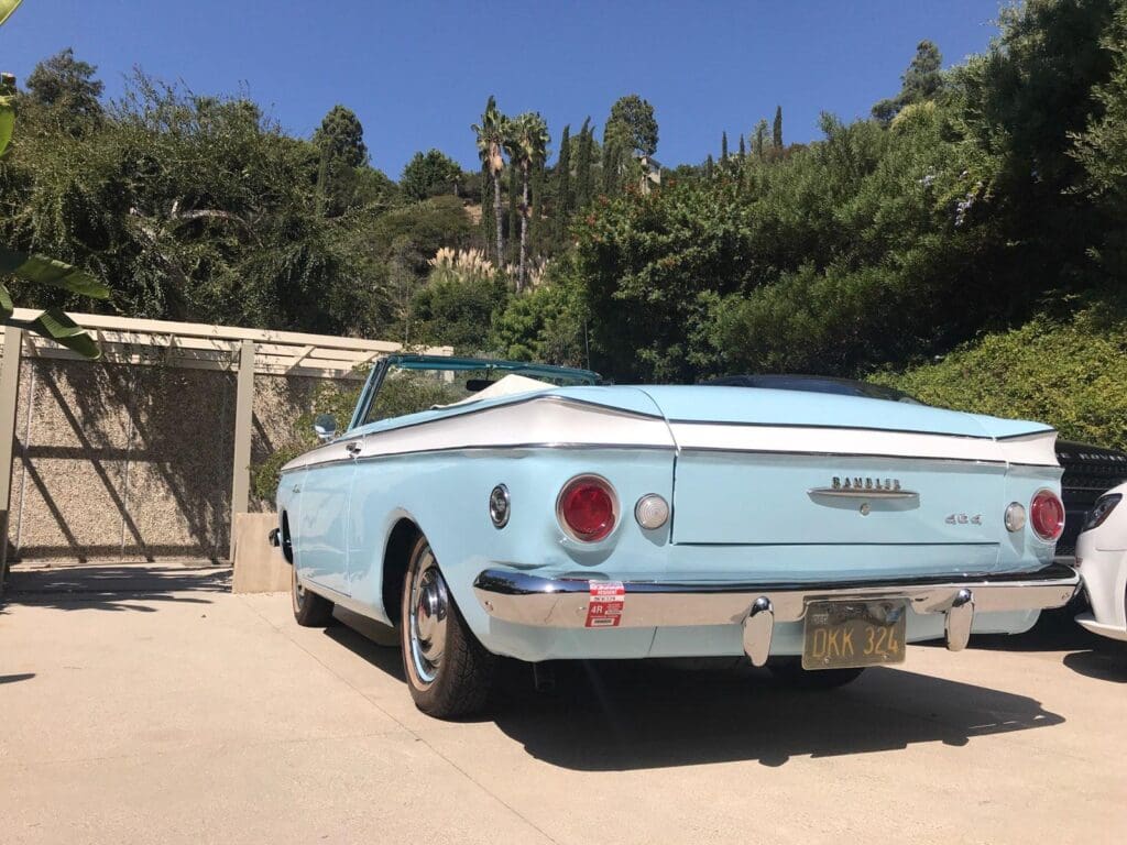 A vintage light blue convertible parked on a sunny driveway with greenery around.