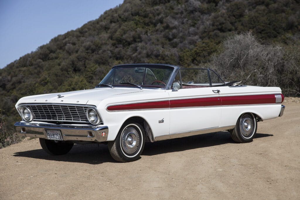 Classic white convertible car parked on a dirt road near a hill.