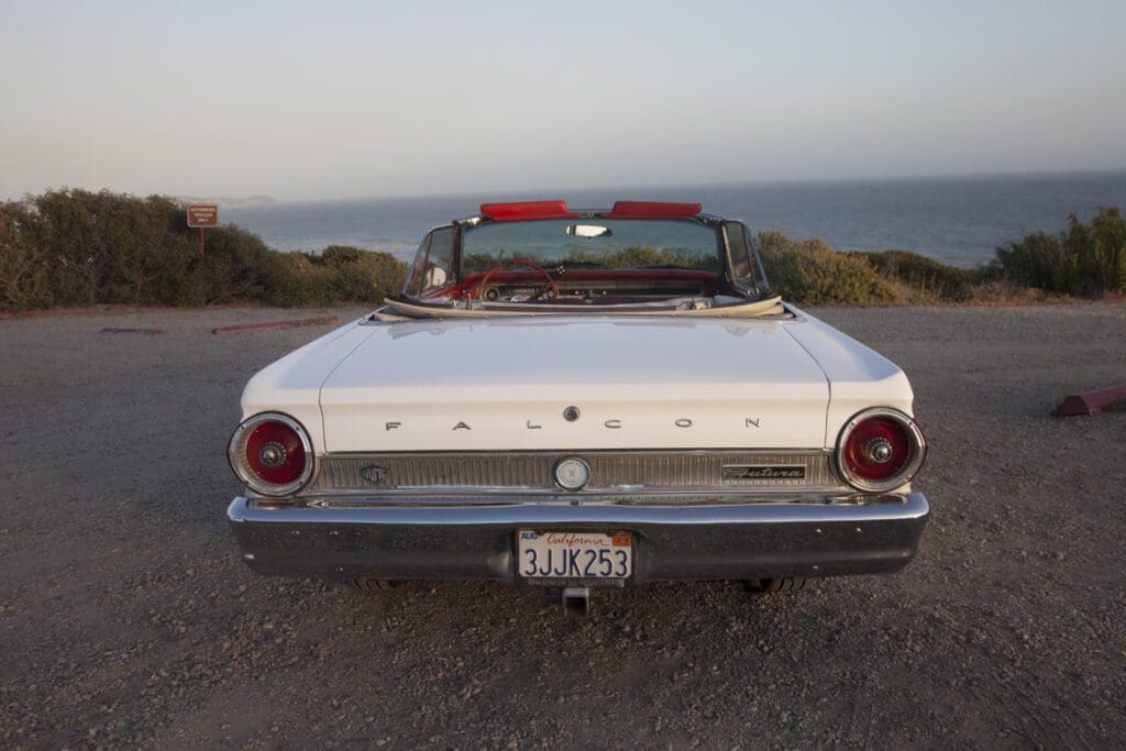 Classic white Ford convertible parked on a coastal road at sunset.