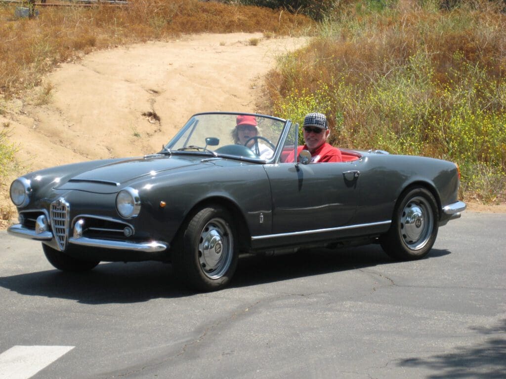 A classic convertible car with two passengers driving on a sunny road.