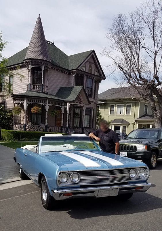 Classic blue convertible parked in front of a Victorian house with a person nearby.