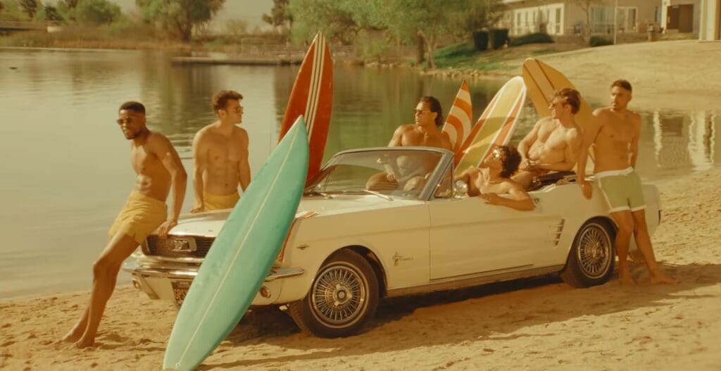 Friends enjoying a sunny beach day with a classic convertible car and surfboards.