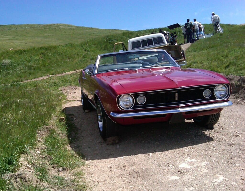 Classic red muscle car parked on a dirt path with a green hill in the background.