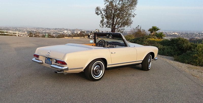 Classic cream convertible car parked on a road with a city view.