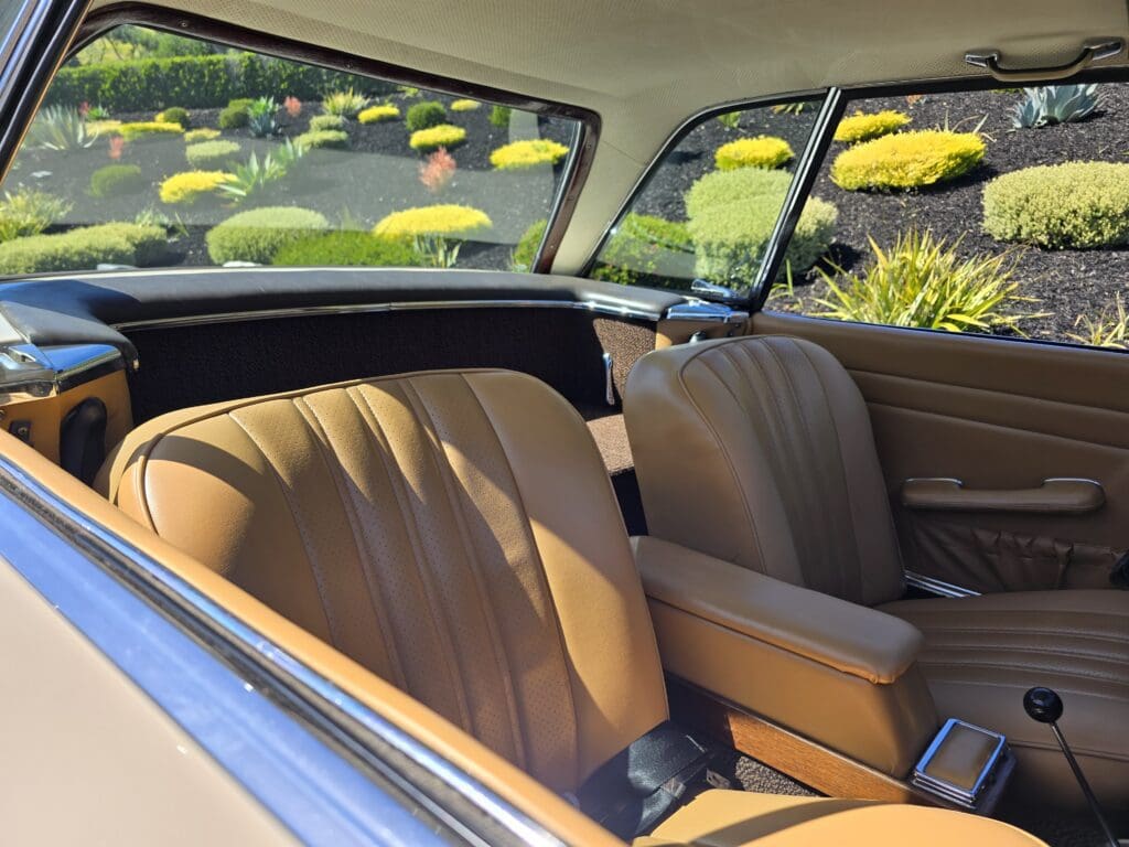 Interior view of a classic car with tan leather seats and lush greenery outside.