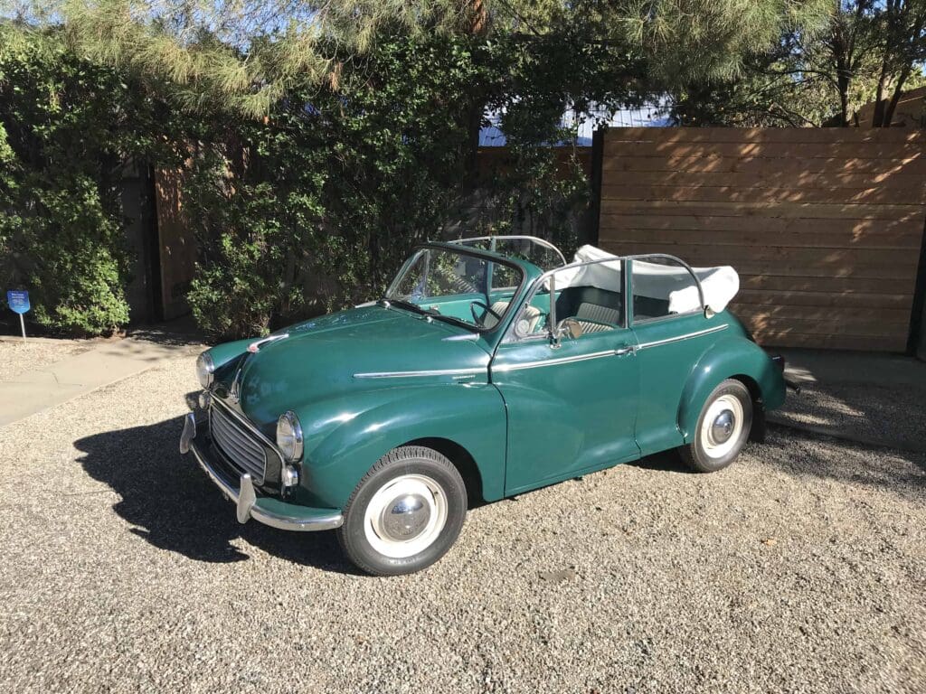 Classic green convertible car parked on gravel under trees.