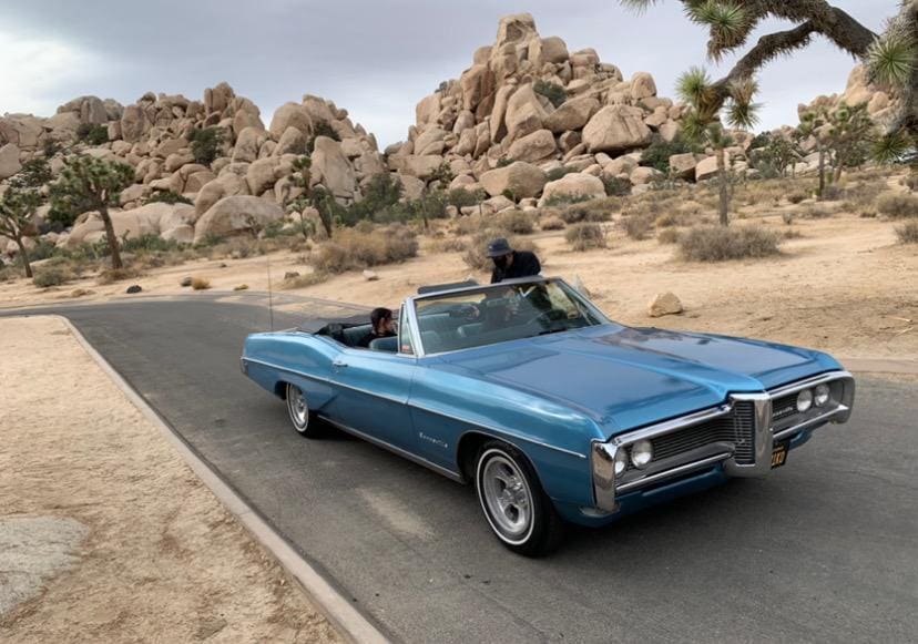 Classic blue convertible car parked on a desert road near rocky formations.