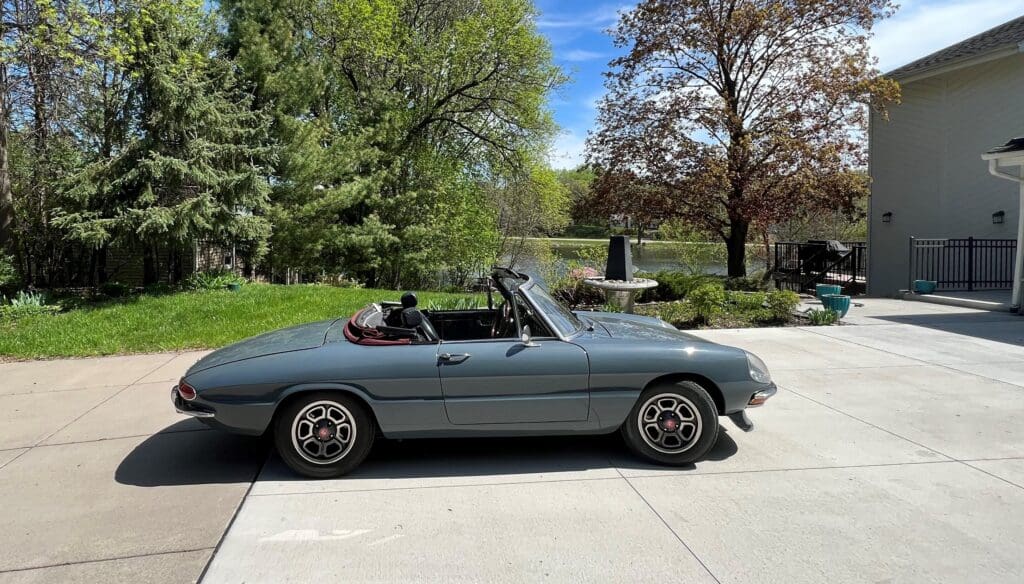 Classic convertible car parked on a sunny driveway with trees and a fountain in the background.