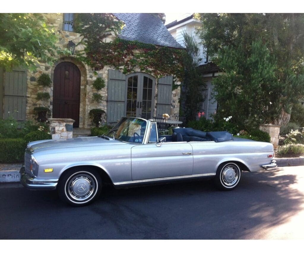 Classic silver convertible parked in front of a rustic stone house.