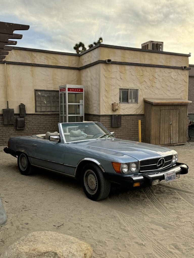 Classic silver convertible car parked near a building with a phone booth.