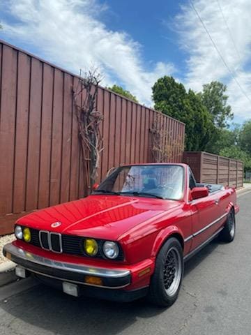 Red classic BMW convertible parked by a wooden fence on a sunny day.