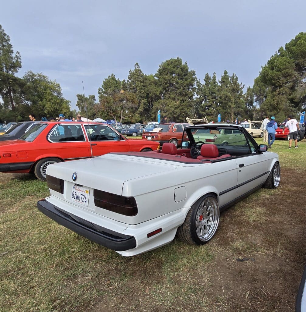 White convertible car displayed at an outdoor car show with red cars in the background.