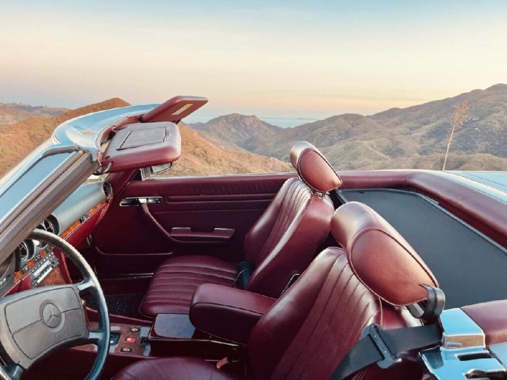 Vintage convertible car with red leather interior parked in a mountainous area at sunset.