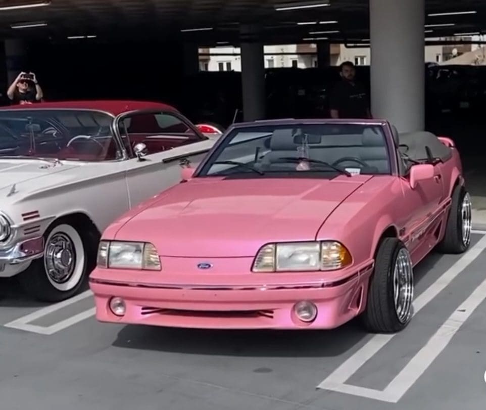 A pink convertible sports car parked in a garage beside another classic car.