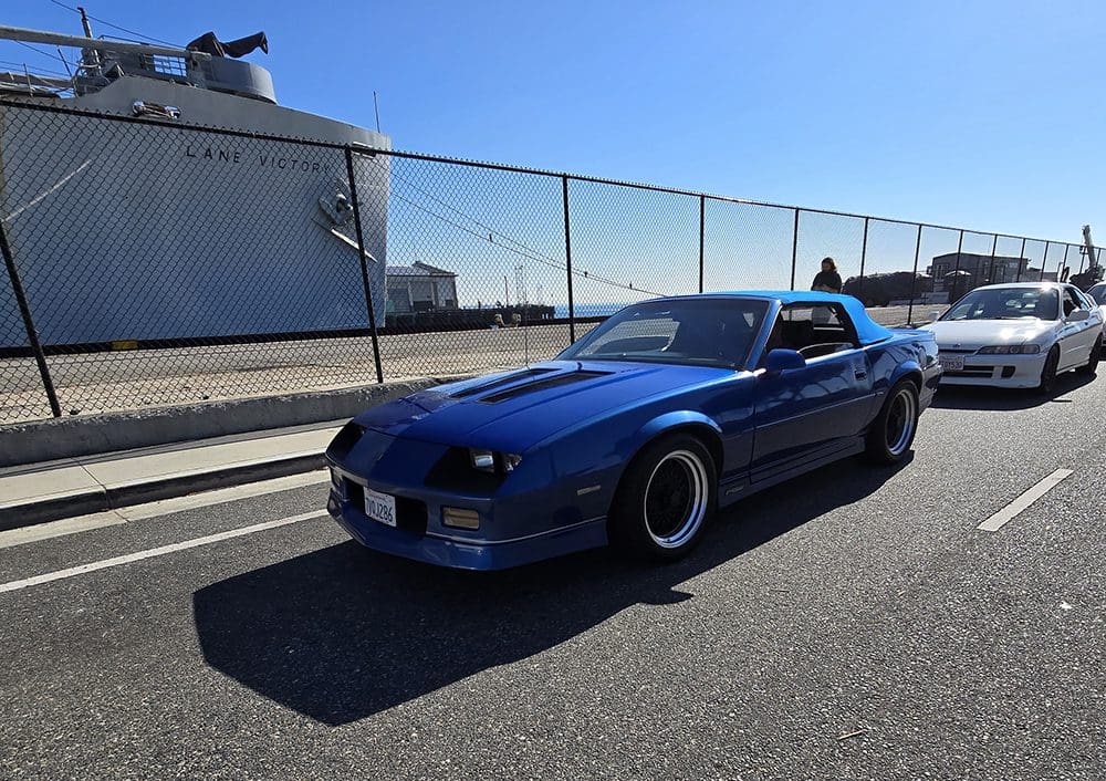 Blue sports car parked near a chain-link fence on a sunny day.