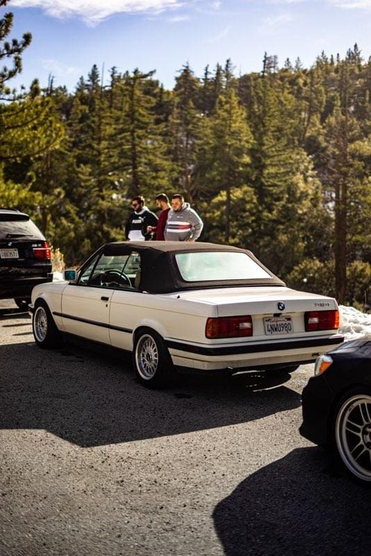 Classic white car parked with luggage on roof in a forest setting.