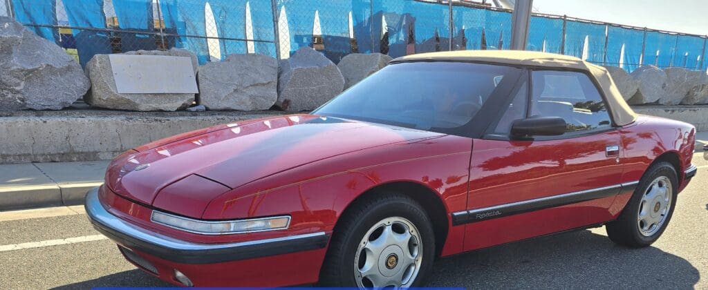 Red vintage sports car parked outdoors near rocks and blue fencing.