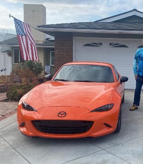 Bright orange Mazda MX-5 Miata parked in a driveway beside a person.
