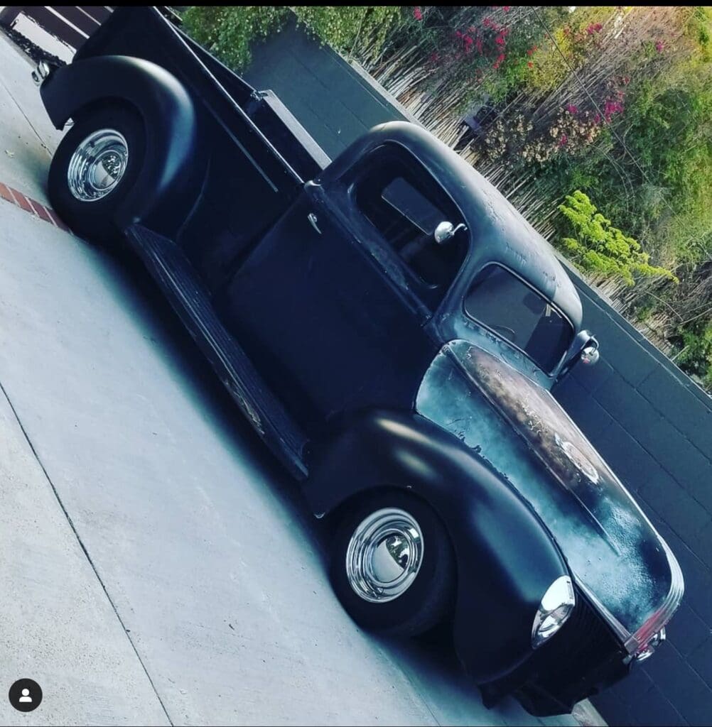 Classic black vintage pickup truck parked on driveway with colorful trees in background.