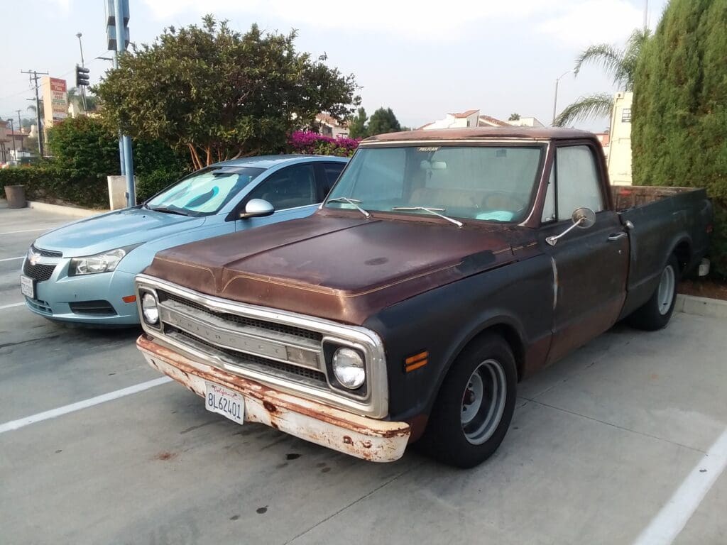 Rusty vintage pickup truck parked beside a blue car in a driveway.