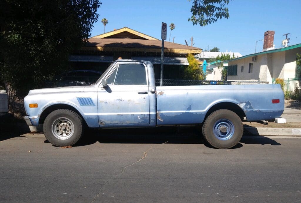 Old blue pickup truck parked on a street under a tree.
