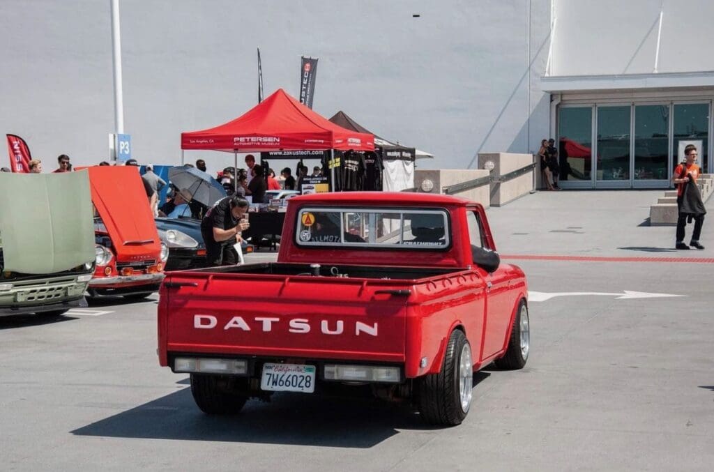 Red Datsun pickup truck parked outdoors near tents and people.