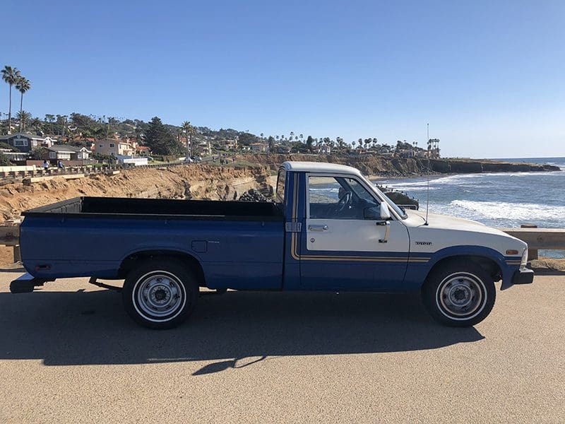 Blue and gray pickup truck parked by the seaside with coastal houses in the background.