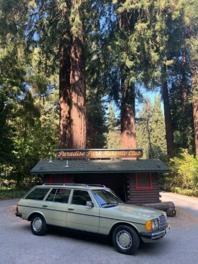 A vintage white station wagon parked in front of a cabin surrounded by tall trees.