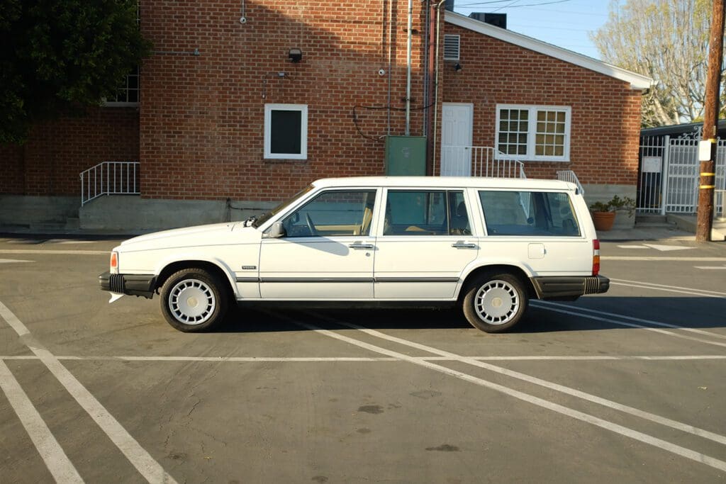 A vintage white station wagon parked in an empty lot beside a brick building.