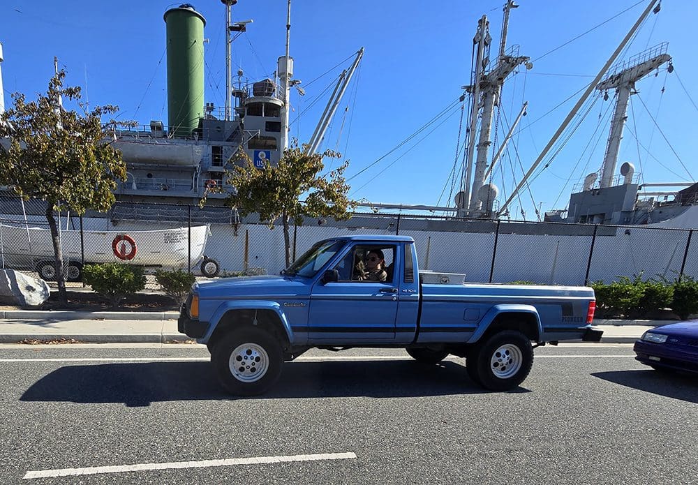 A blue pickup truck parked on a street near a docked ship.